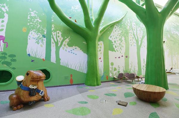 A children's hospital features a room with a nature theme with green trees, colorful wall murals, and a wooden frog