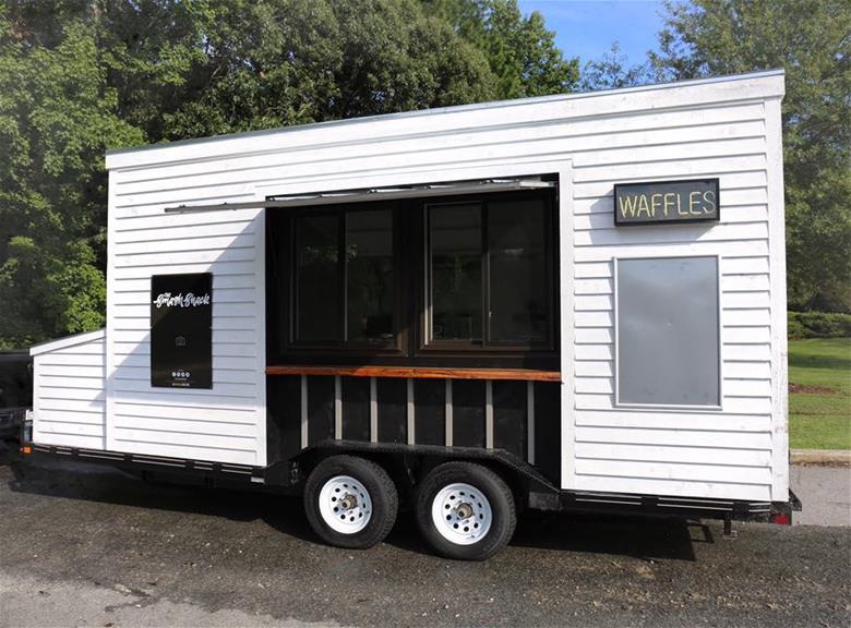 A food truck with white paneling has a sign displayed that lights up to spell the word "Waffles"