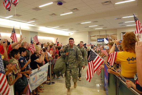 Military members walk through a crowded airport with people waving flags and holding signs