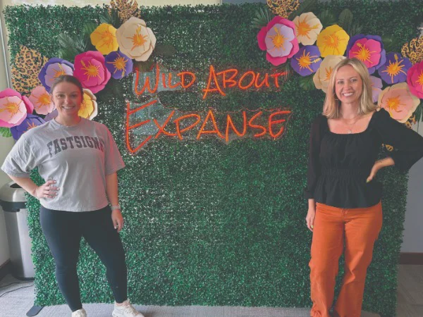 two women smiling in front of an LED sign that reads "Wild About Expanse"